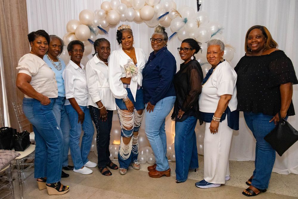 Group of women posing together at a celebration event, wearing blue jeans and white or dark tops with balloon decorations.