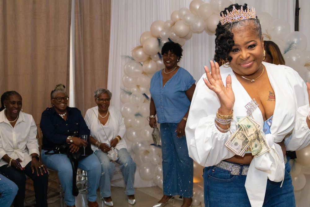 Woman in white outfit and tiara holds cash at a festive celebration with balloon backdrop and seated guests.