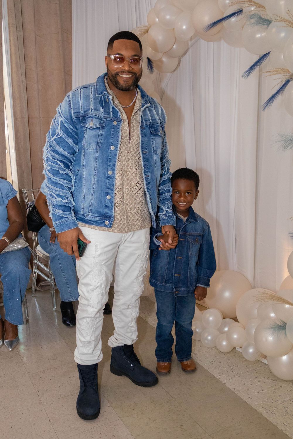 Man and boy in matching denim jackets pose together at a balloon-decorated event.