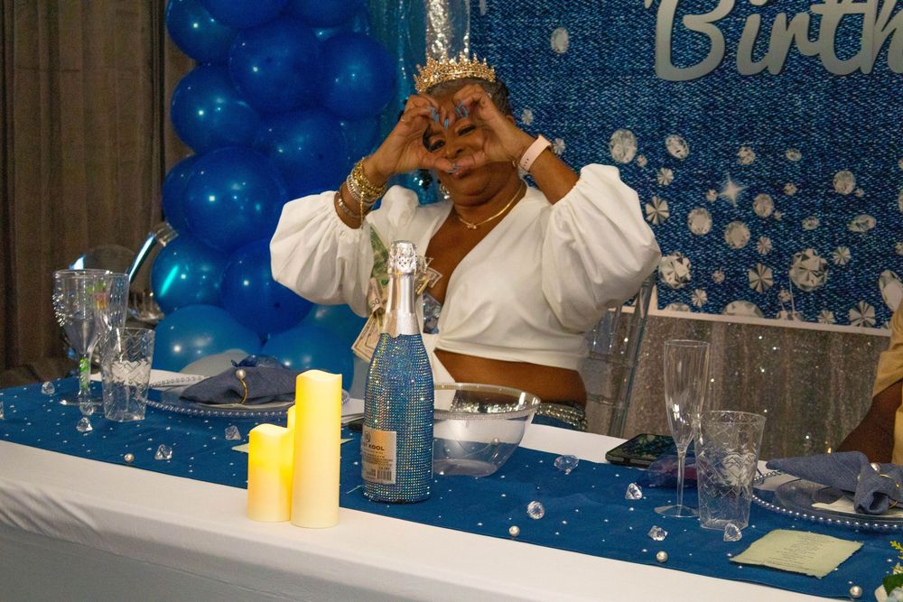 Woman making heart hand gesture at blue birthday party table with balloon decorations and champagne bottle.