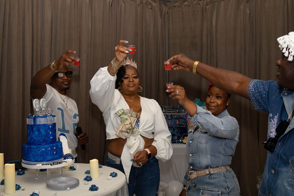 Group celebrating birthday party with blue cake, raising glasses in toast while woman holds cash money.