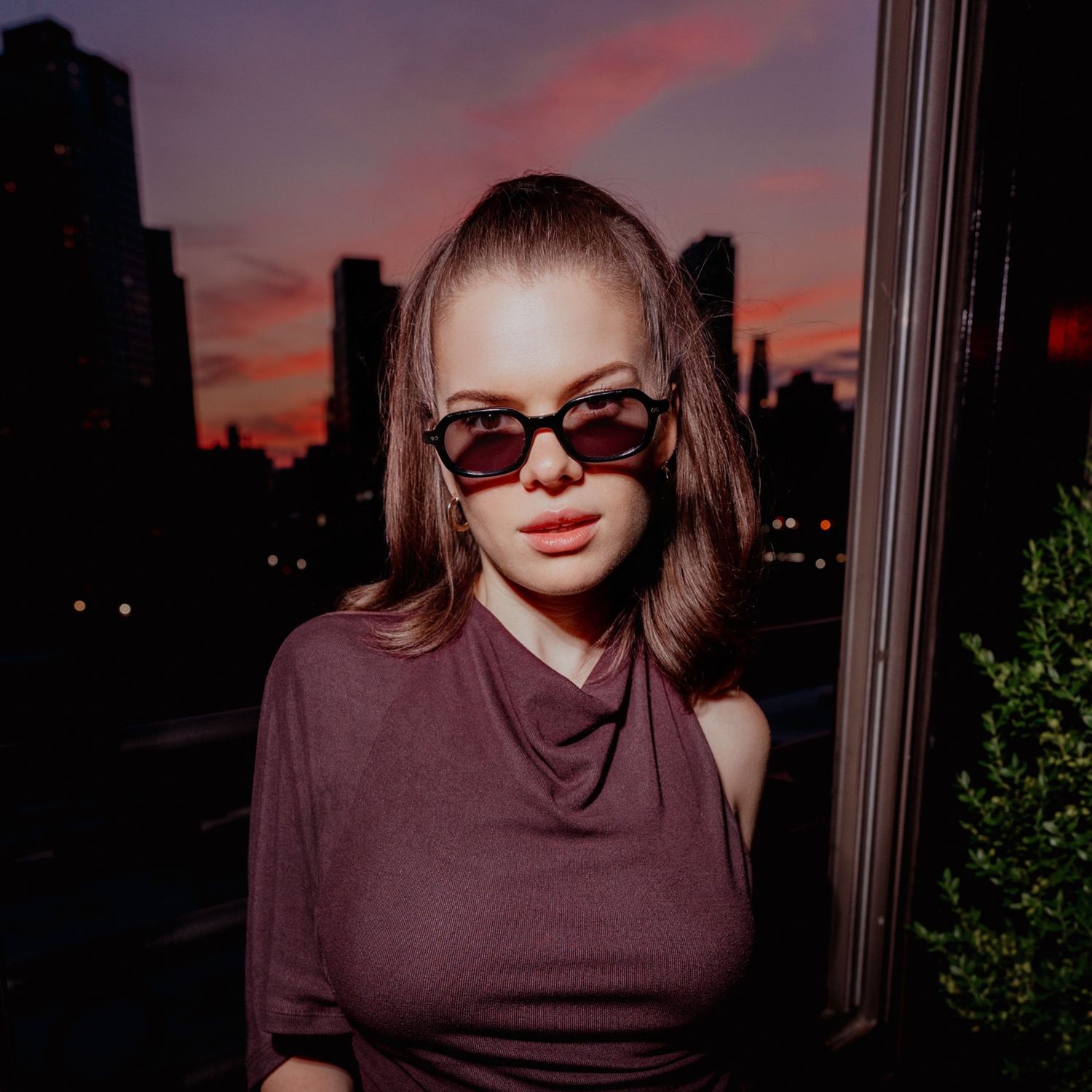 Young woman in sunglasses poses against city skyline at sunset dusk.