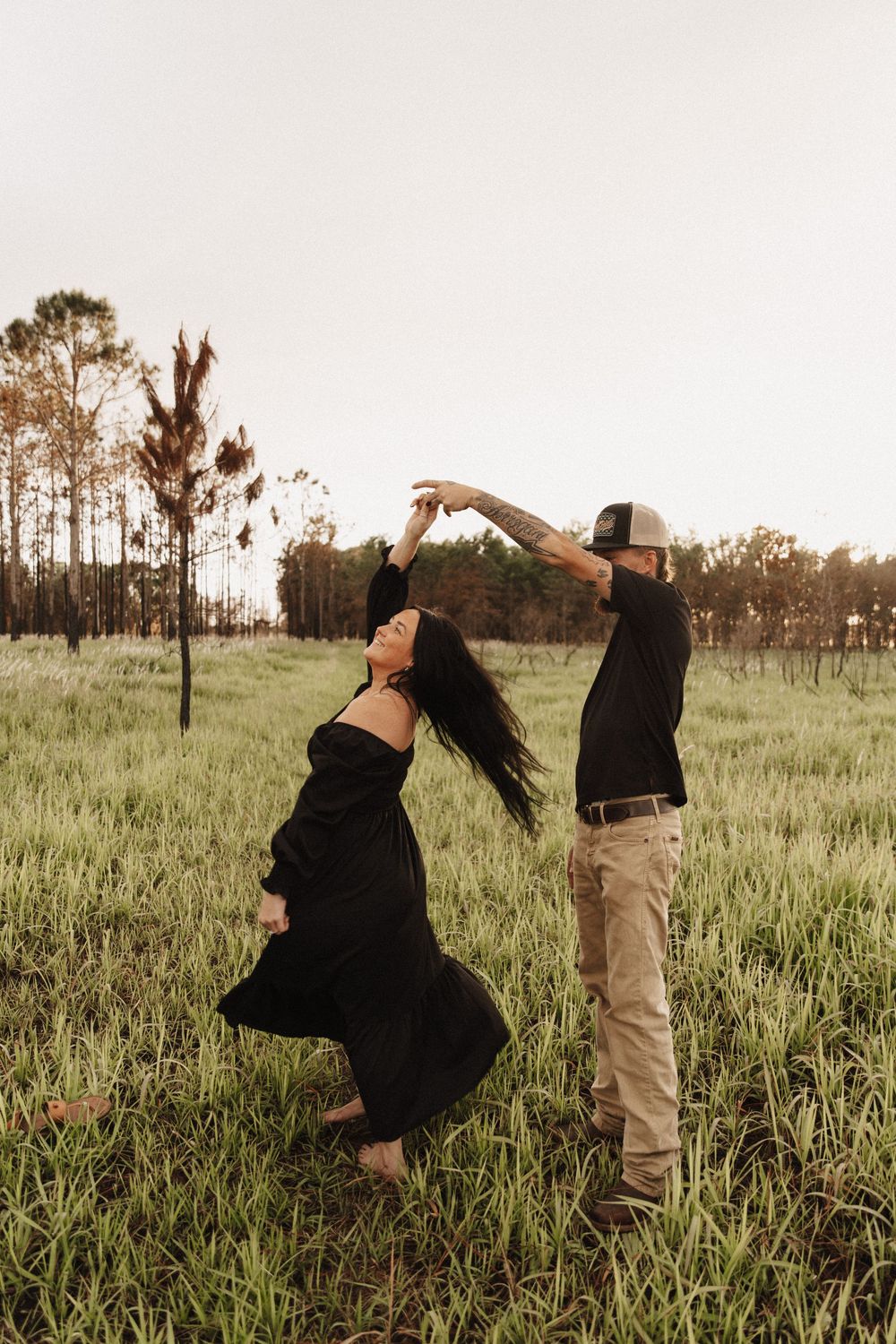 Two people dance joyfully through a grassy meadow at sunset with pine trees on the horizon.