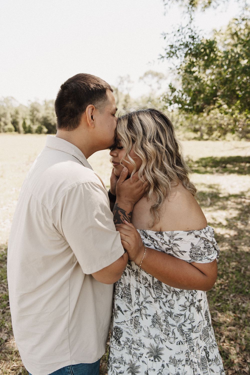 A romantic couple embrace and kiss while standing in a sunlit outdoor setting wearing white and floral clothing.
