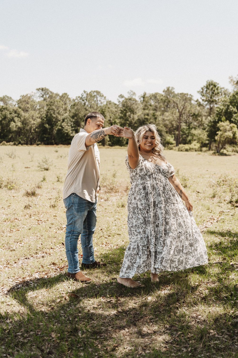 Two people dance together playfully in a sunny outdoor field surrounded by trees and grass.