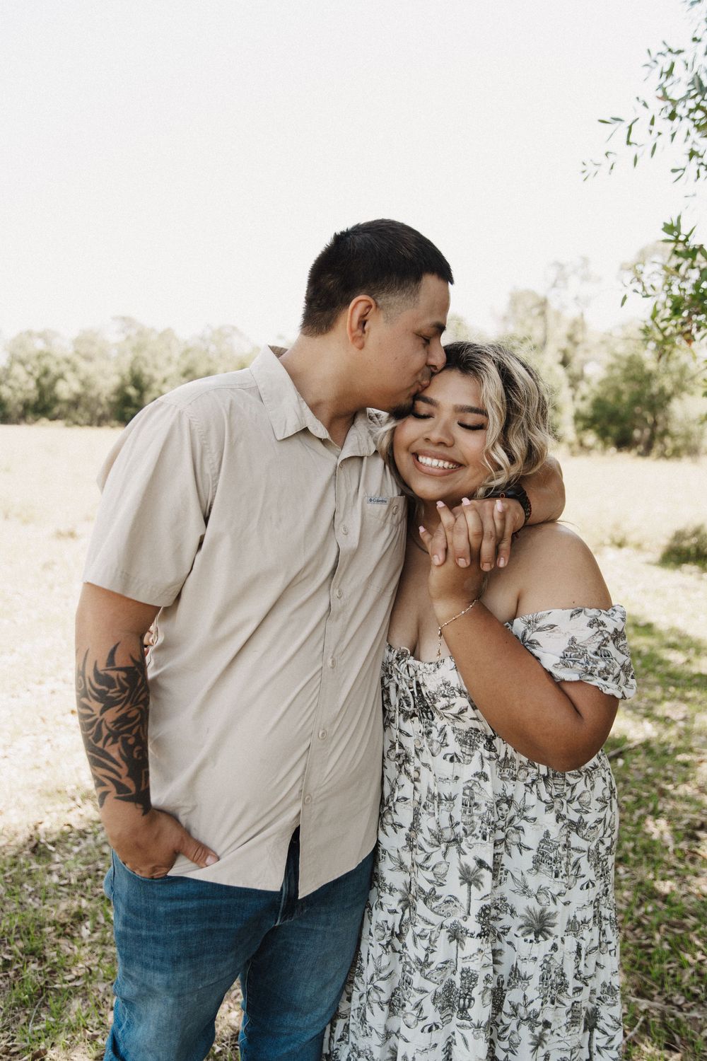 A romantic couple shares an intimate moment outdoors, with her wearing a floral dress and him in a beige shirt.