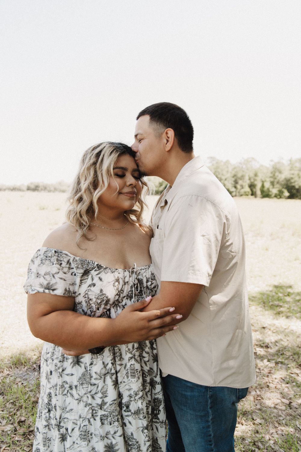 A couple shares romantic moments together in an outdoor field setting wearing white and floral outfits.