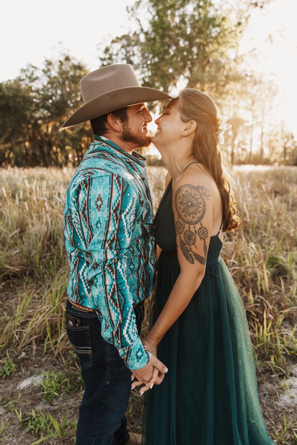 A couple sharing a romantic sunset kiss in a field, with him in a turquoise western shirt and her in an emerald dress.