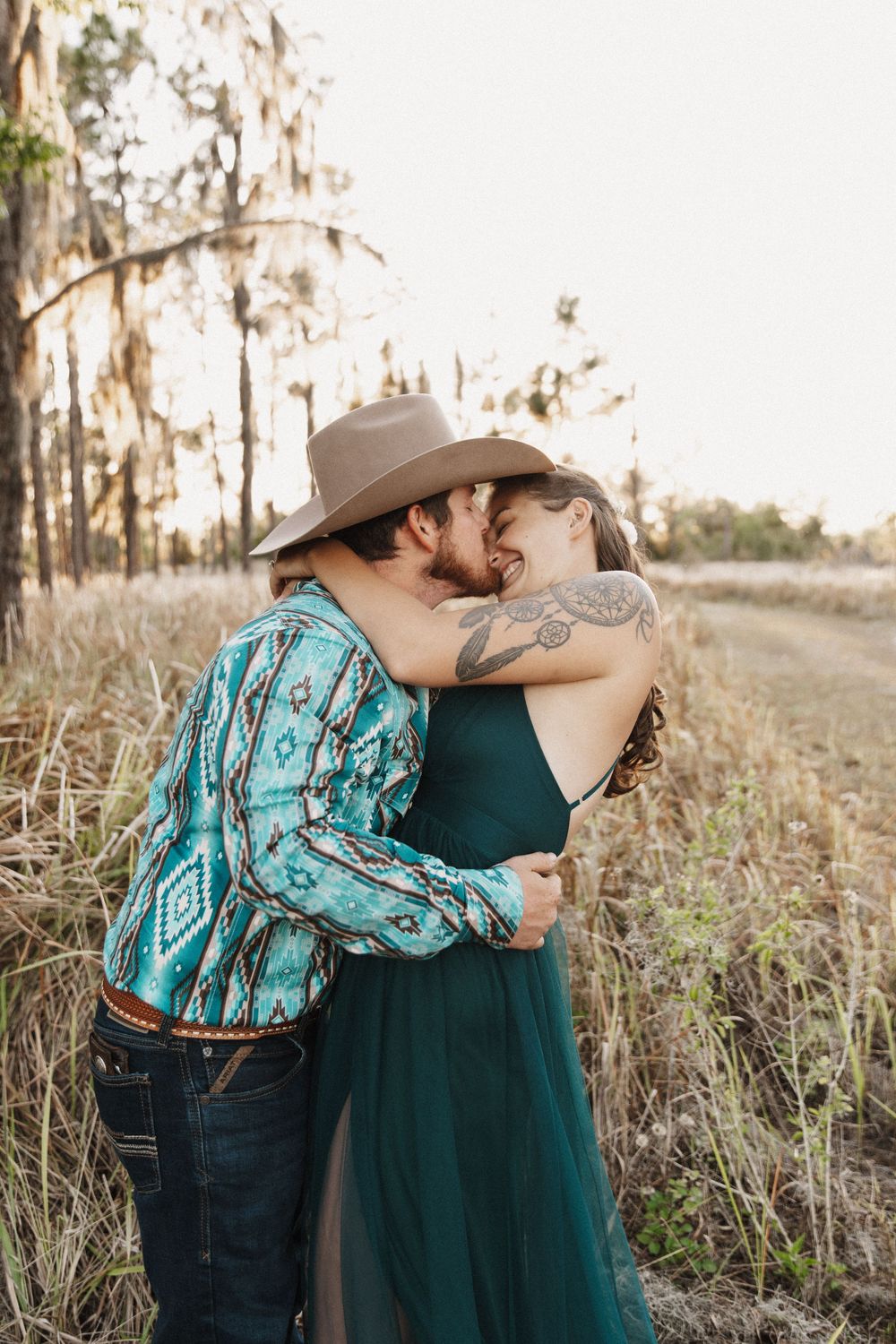 A couple in western attire shares romantic moments in a rustic field at sunset.