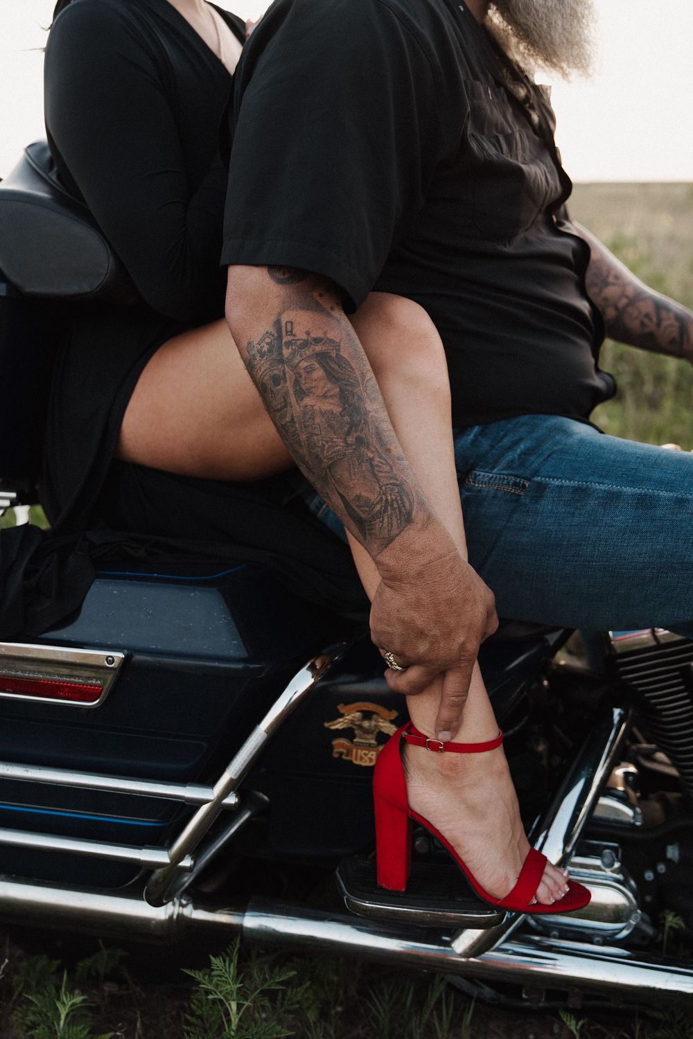 A person wearing jeans sits on a motorcycle, showing a close-up view of their arm tattoo and red heels.