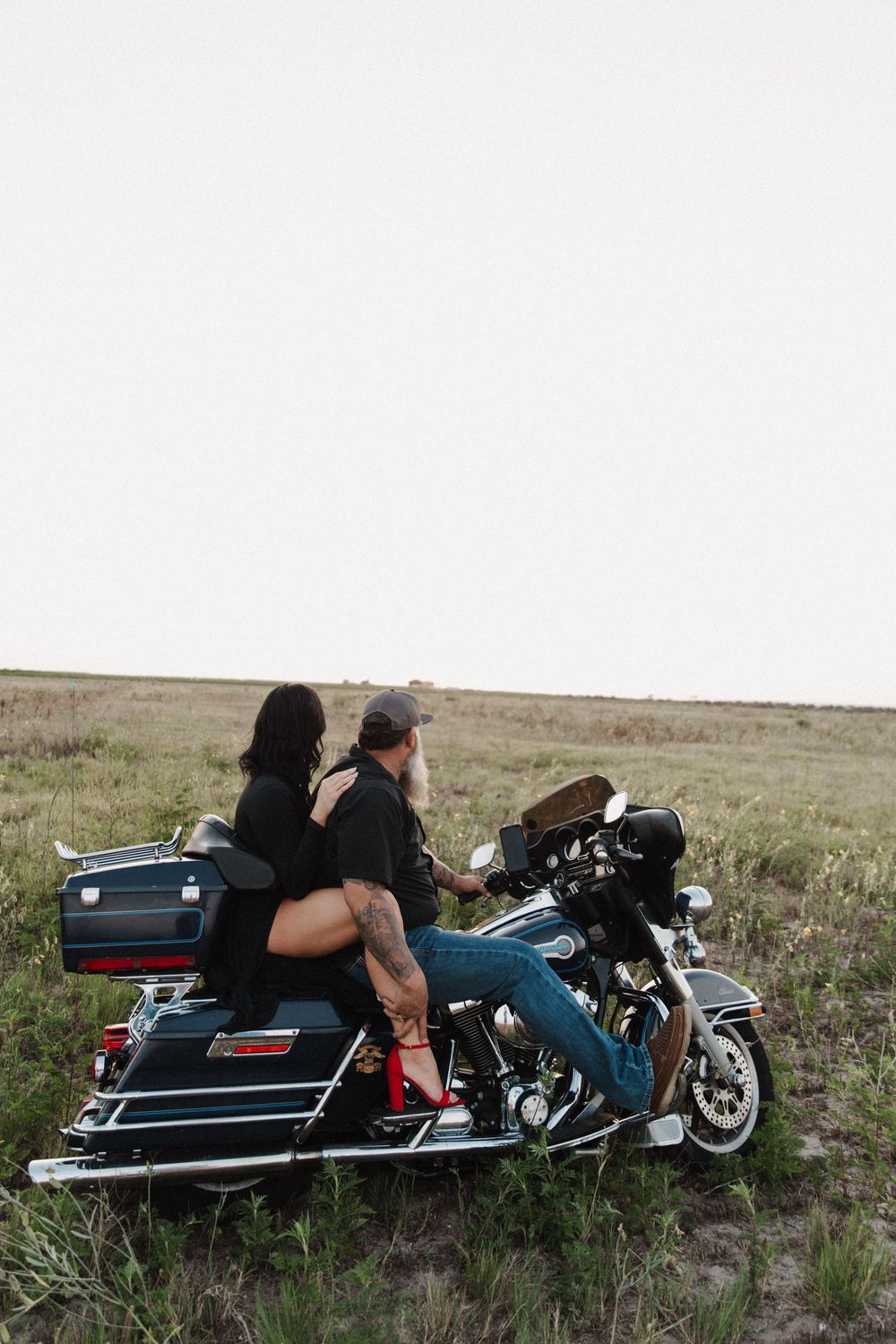 Two people sitting on a parked Harley Davidson motorcycle in an open field at dusk.