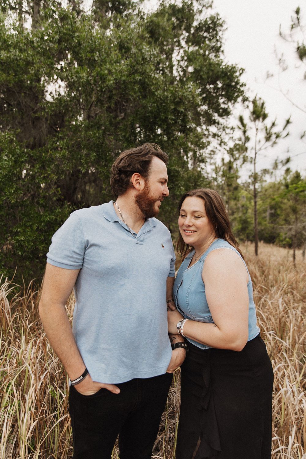 A couple in matching blue shirts and black pants share intimate moments while posing in front of tall grass and trees.