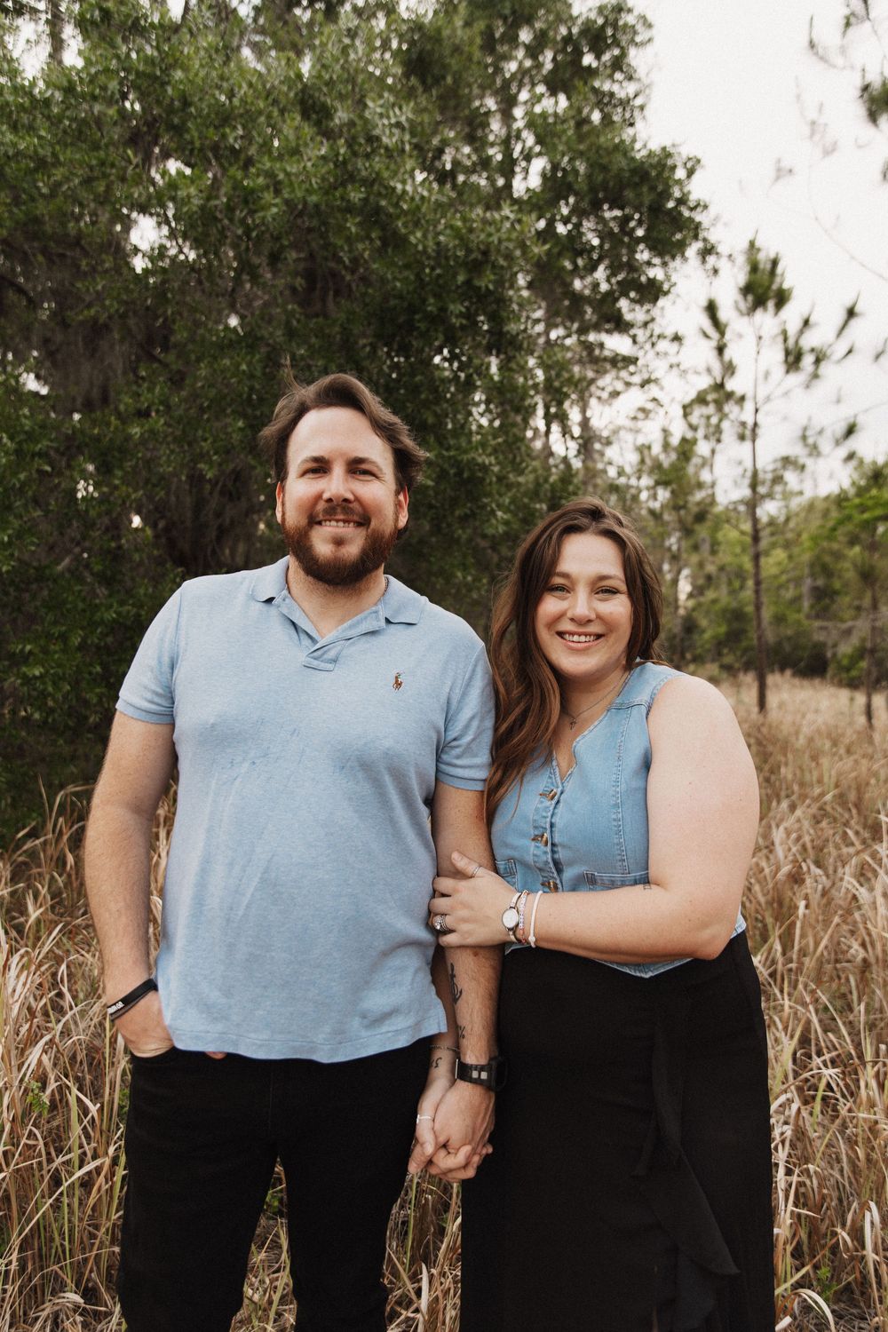 A couple stands together in front of trees and tall grass during a natural outdoor portrait session.