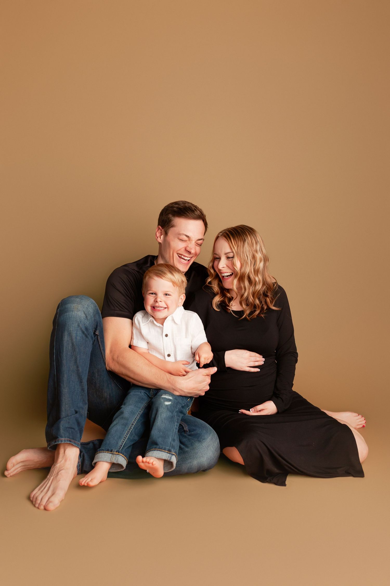 Expecting family of three sitting on a tan studio floor, father holding toddler son beside pregnant mother in black dress.