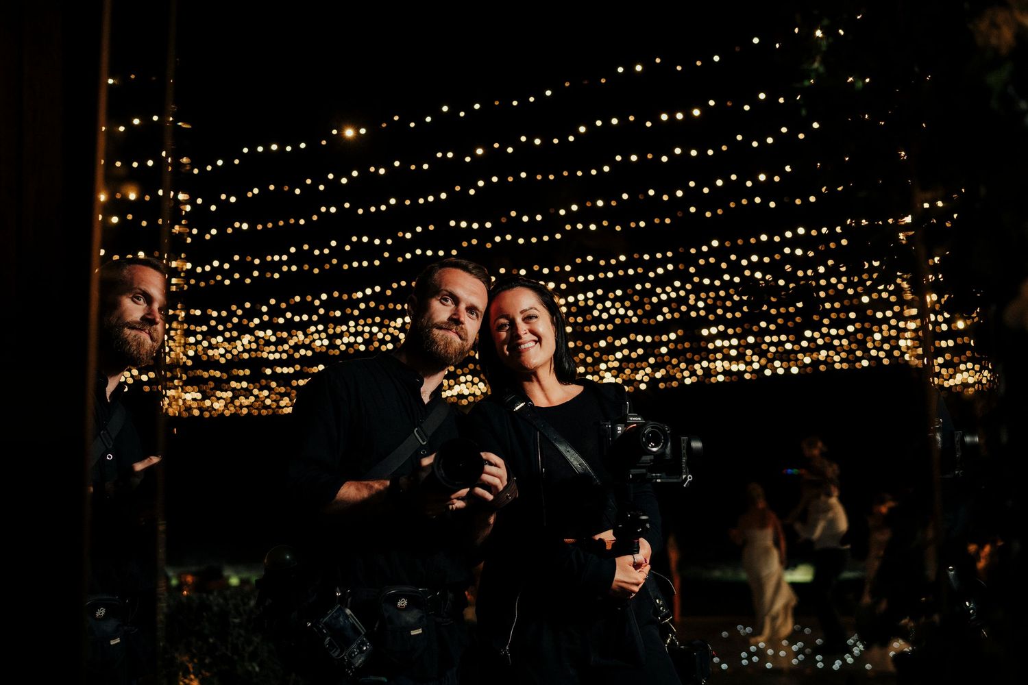 Professional photography duo poses with cameras against grey background in black and white portrait.