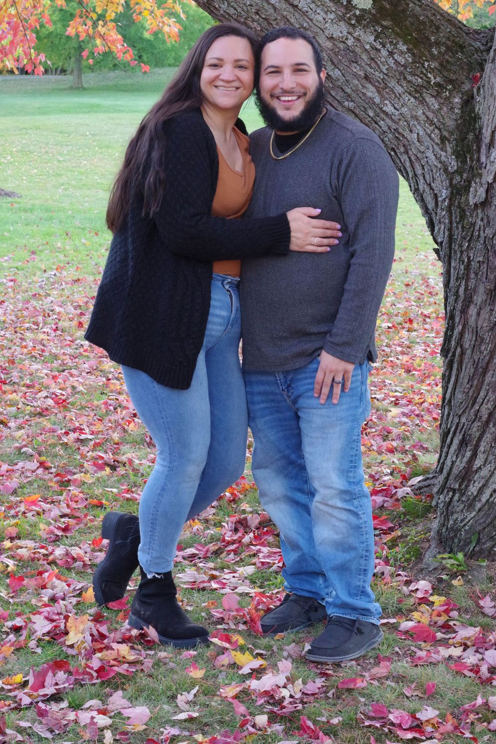 A couple poses together against a tree with colorful autumn foliage in the background.
