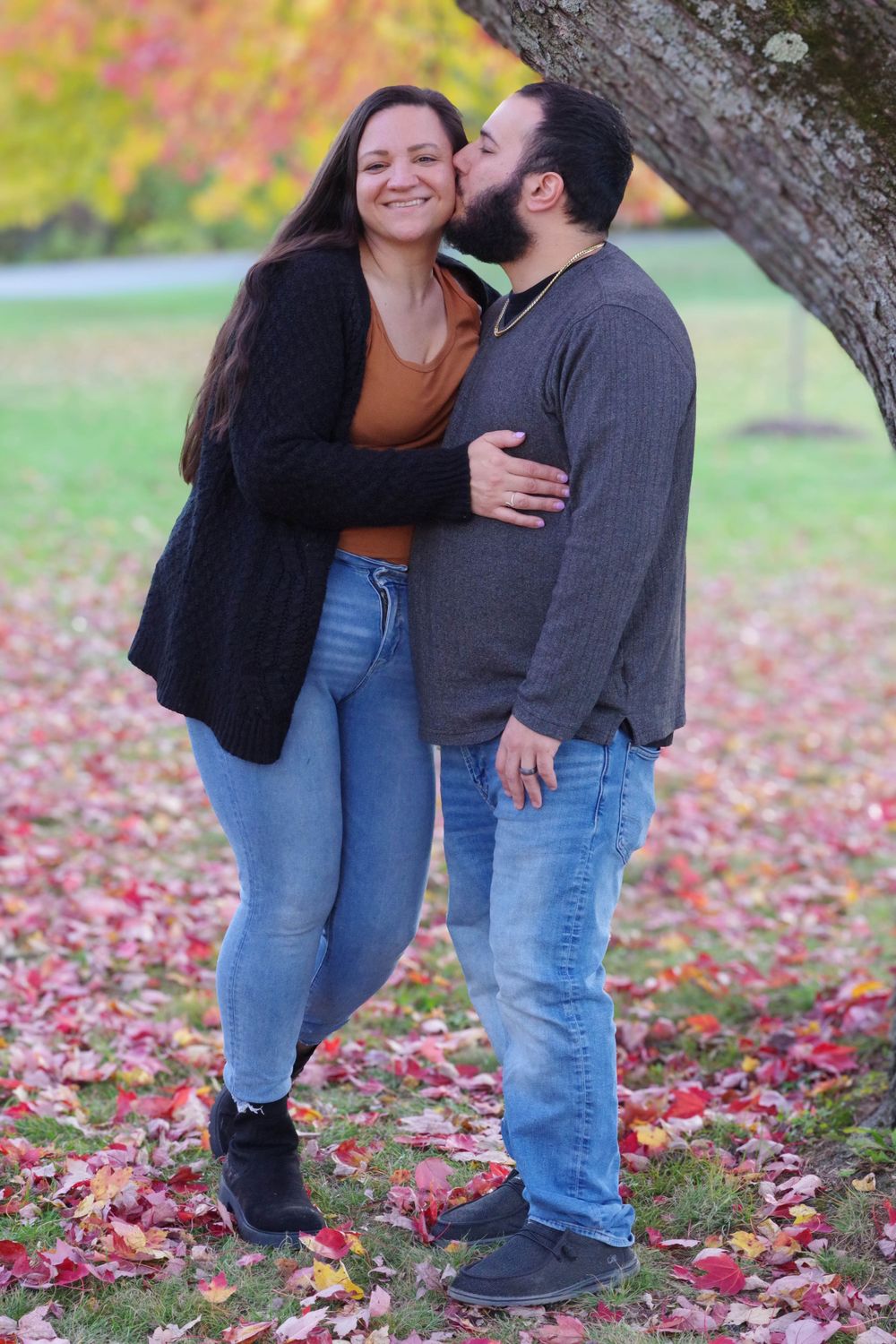 A couple embraces under a tree with red fallen leaves scattered on the ground.
