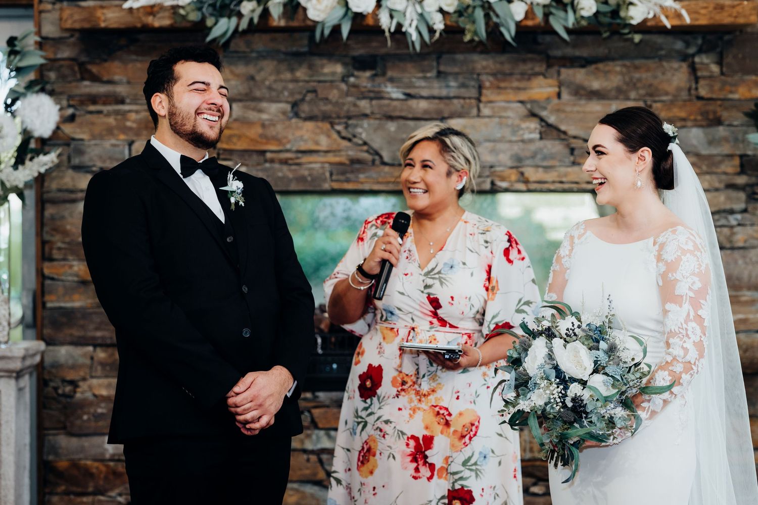 Group wears elegant attire at wedding celebration against rustic stone wall backdrop with white florals and decor.