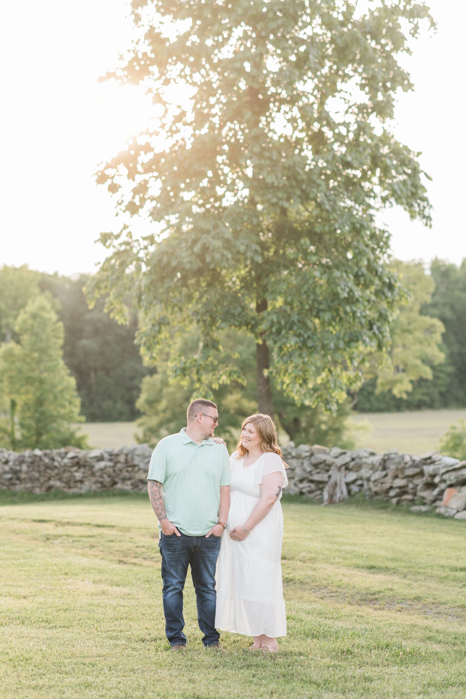 A couple holds hands and gazes at each other in a sunny green field with a stone wall backdrop.