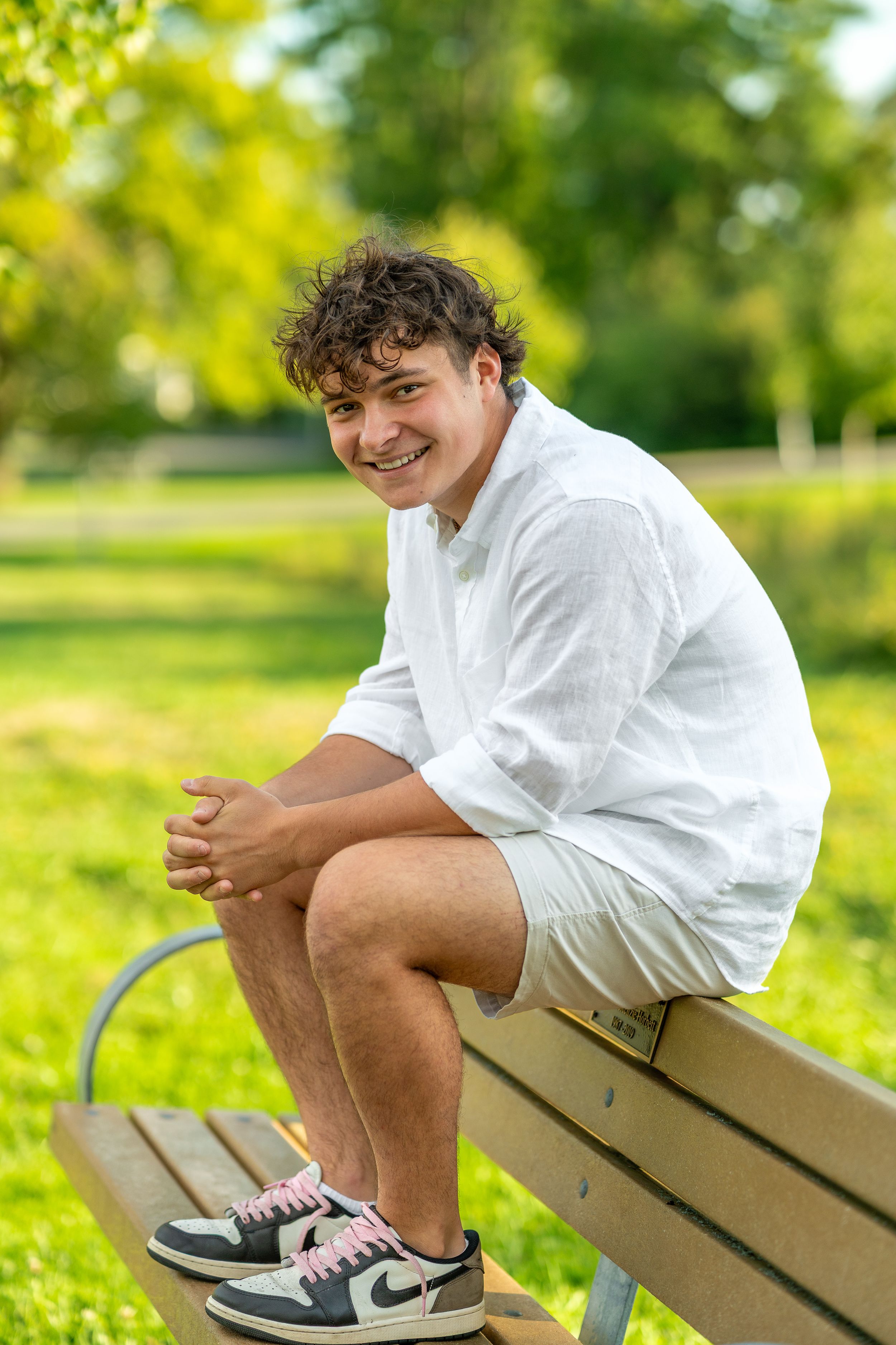 Person in white shirt and shorts sits on wooden bench in sunny park with green grass and trees.