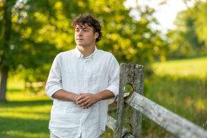 A person in a white button-down shirt stands outdoors in a sunny field with trees and natural lighting.
