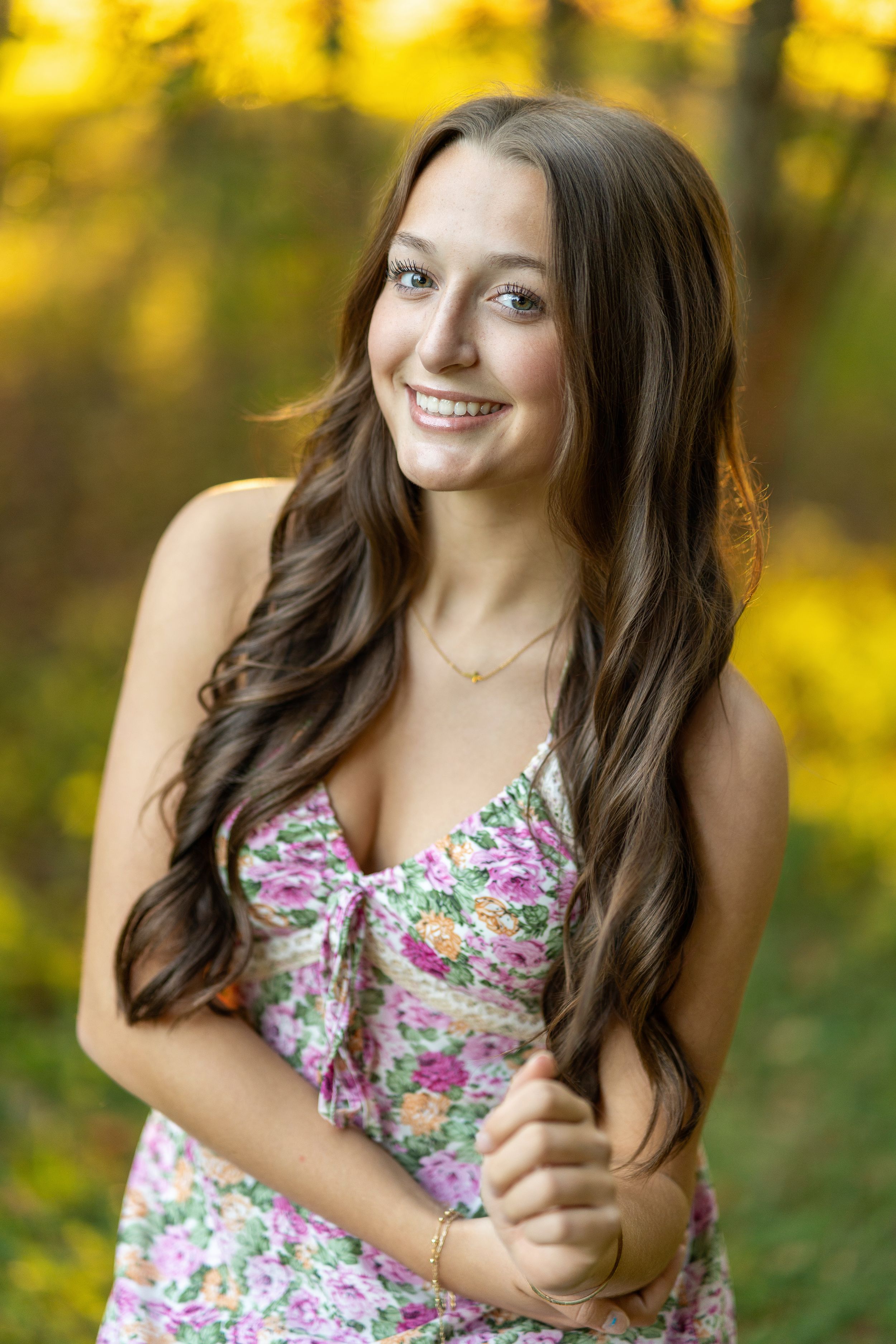 A person in a floral dress poses outdoors with autumn foliage creating a warm yellow backdrop.