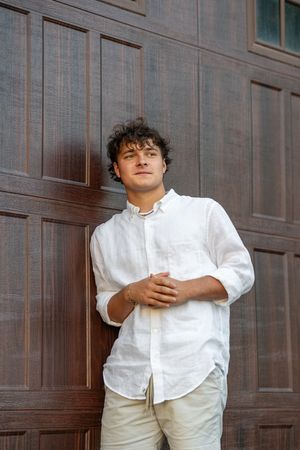 Portrait of someone in a casual white button-down shirt and khaki pants leaning against a wooden paneled wall.