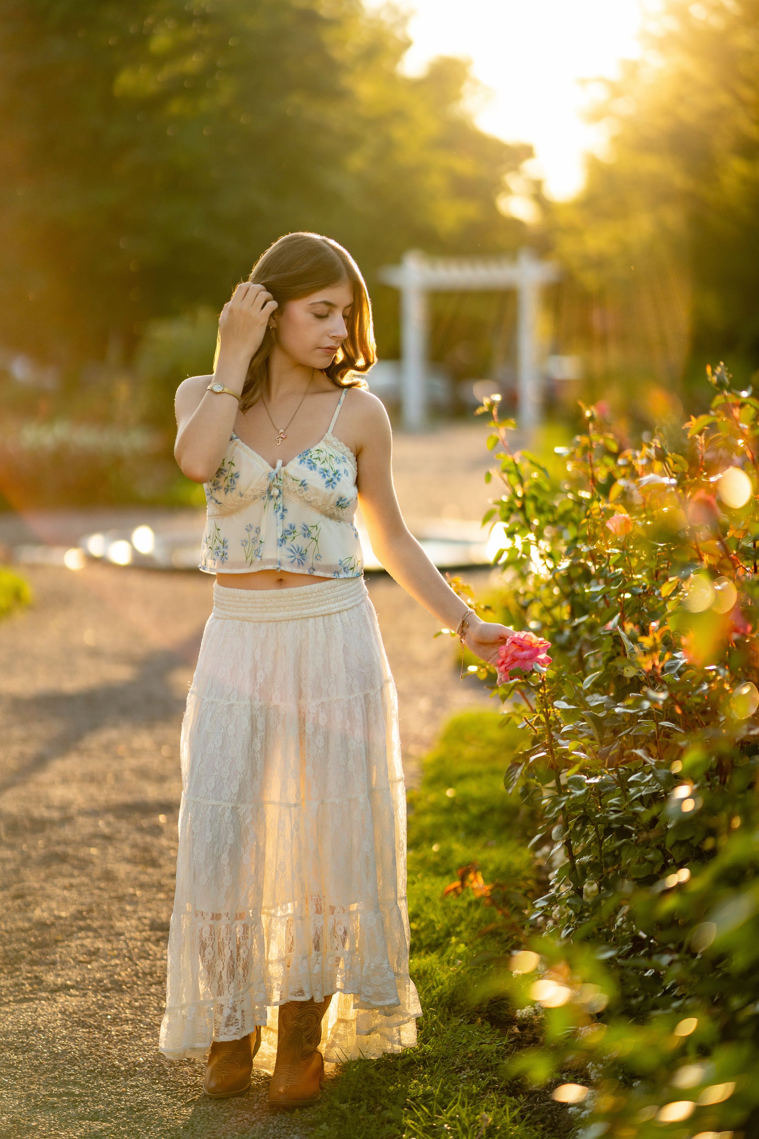 A woman in a floral crop top and white maxi skirt stands by blooming roses at sunset in a garden path.