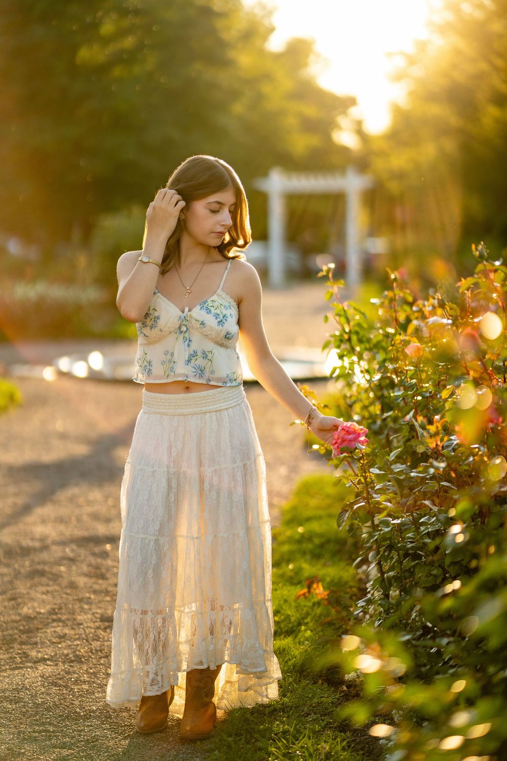A woman in a floral crop top and white maxi skirt stands by blooming roses at sunset in a garden path.