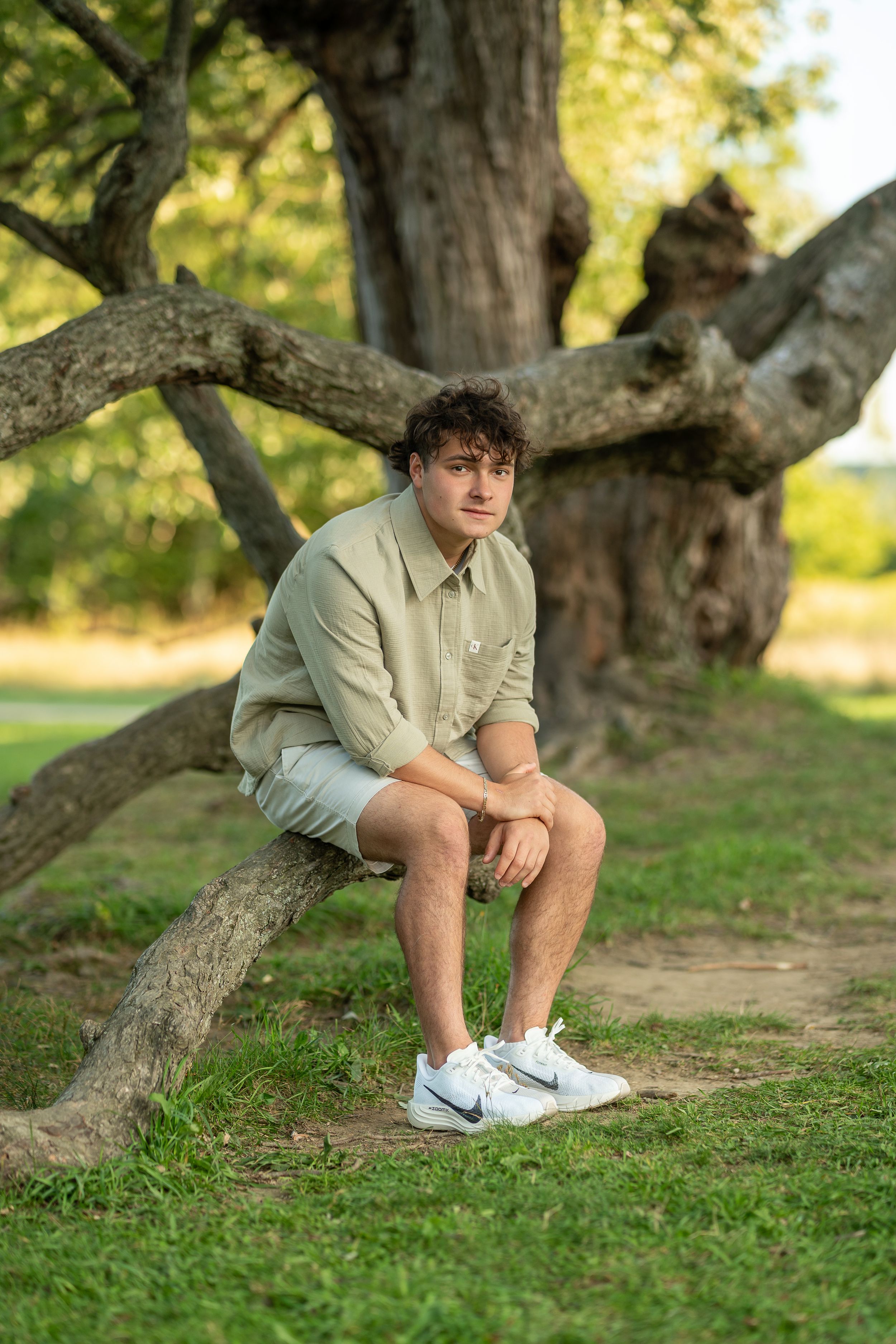 Person in beige outfit poses on grassy ground near gnarled tree trunk in outdoor setting.