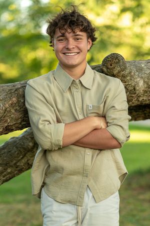 Casual outdoor portrait with someone in a beige button-down shirt leaning against a rustic wooden fence.