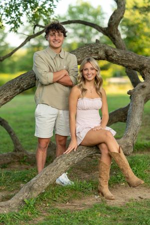 A couple poses together on a curved tree branch in a sunny outdoor setting wearing summer casual attire.