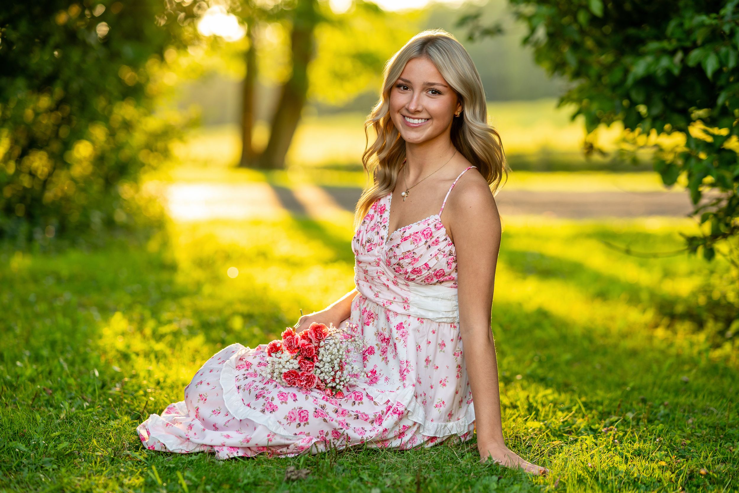 A woman in a floral sundress sits on grass during golden hour with sunlit trees in the background.
