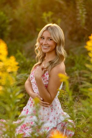 A woman in a floral dress poses gracefully in a sunlit meadow with golden wildflowers and natural greenery.