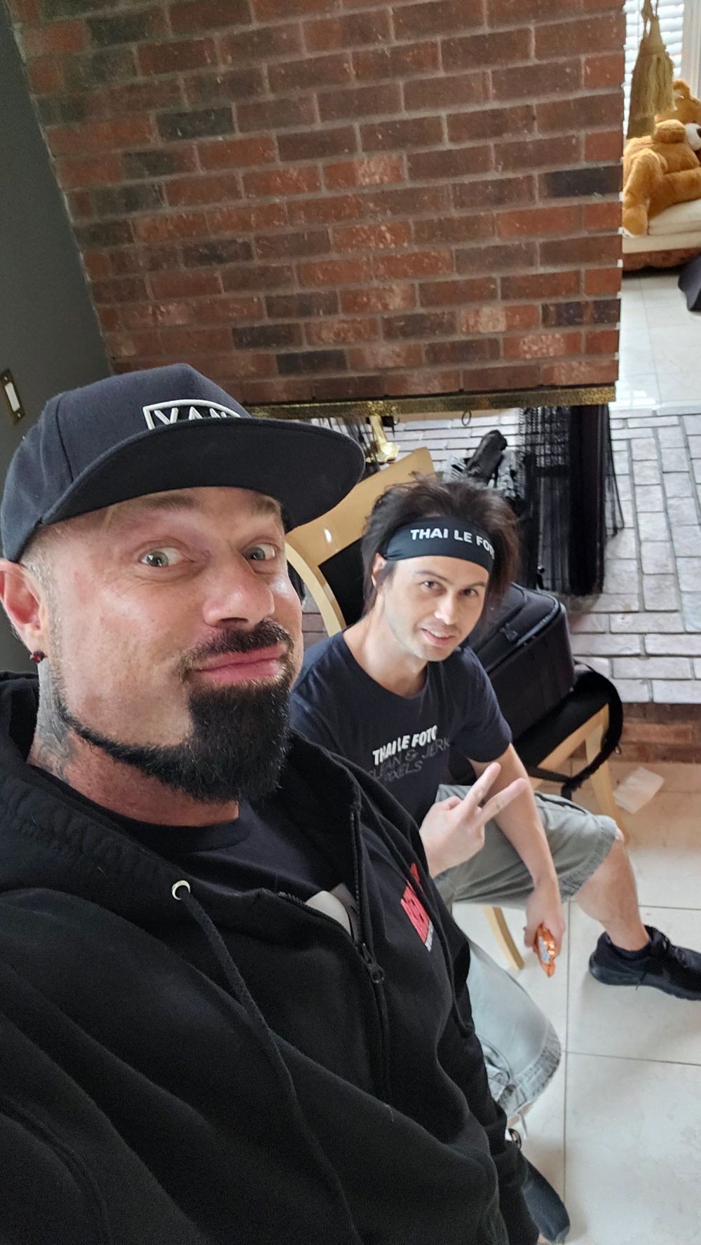 Two people in black hoodies and beanies taking a selfie indoors next to a brick wall.
