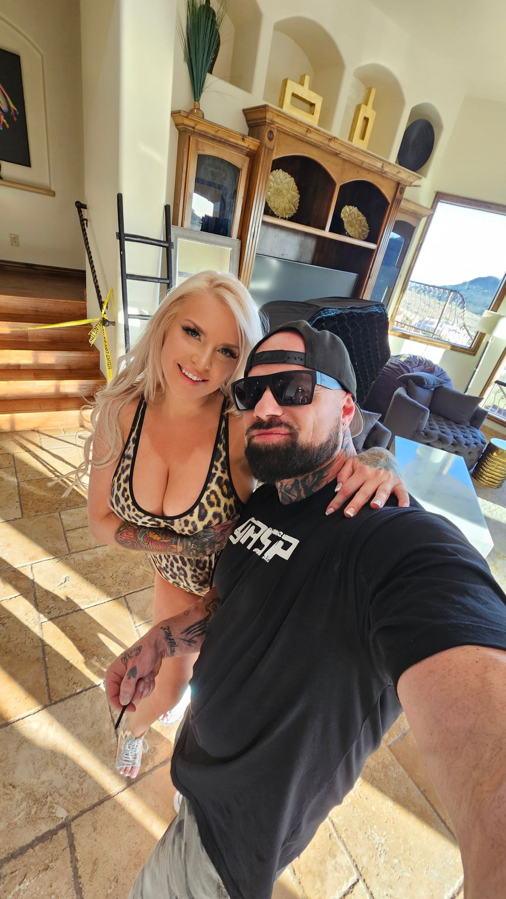 Two people pose for a selfie in a home interior with stone tiles and wooden shelving in the background.