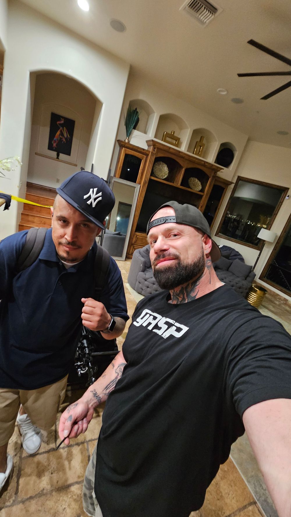 Two men pose for a casual selfie indoors, wearing casual streetwear including a Yankees cap and t-shirt with tattoos visible.