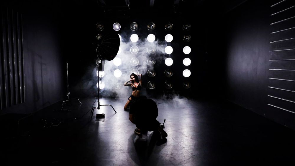 A silhouetted figure crouches in front of bright studio lights creating a dramatic backlit effect in a dark room.