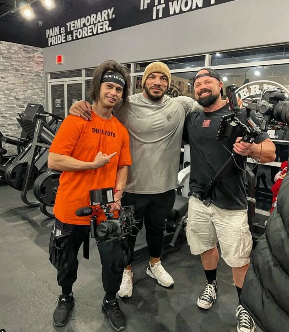 Three people posing together in a gym with exercise equipment and brick walls visible in the background.