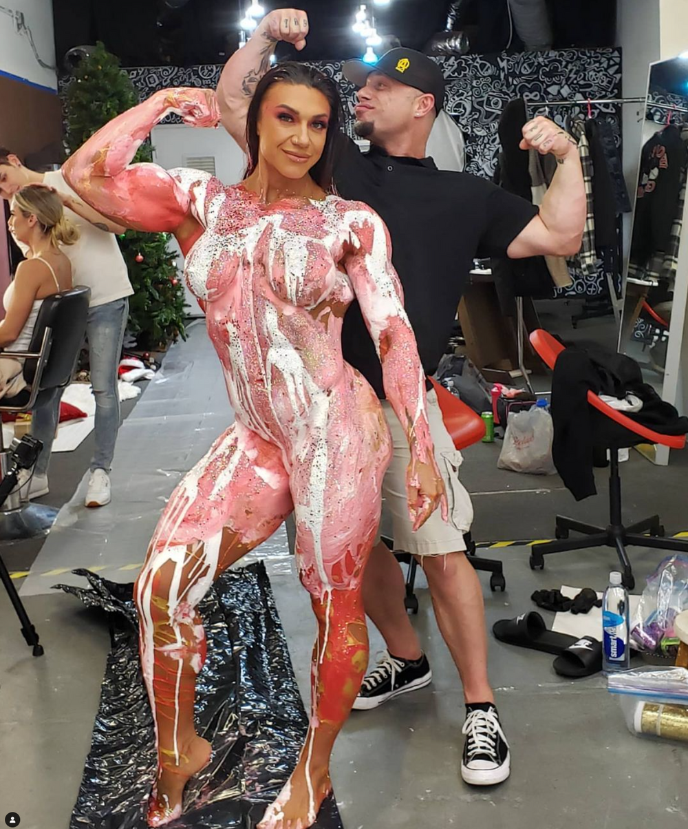 Body painted performer strikes a pose while showing off detailed pink and white muscular anatomy artwork in a studio setting.
