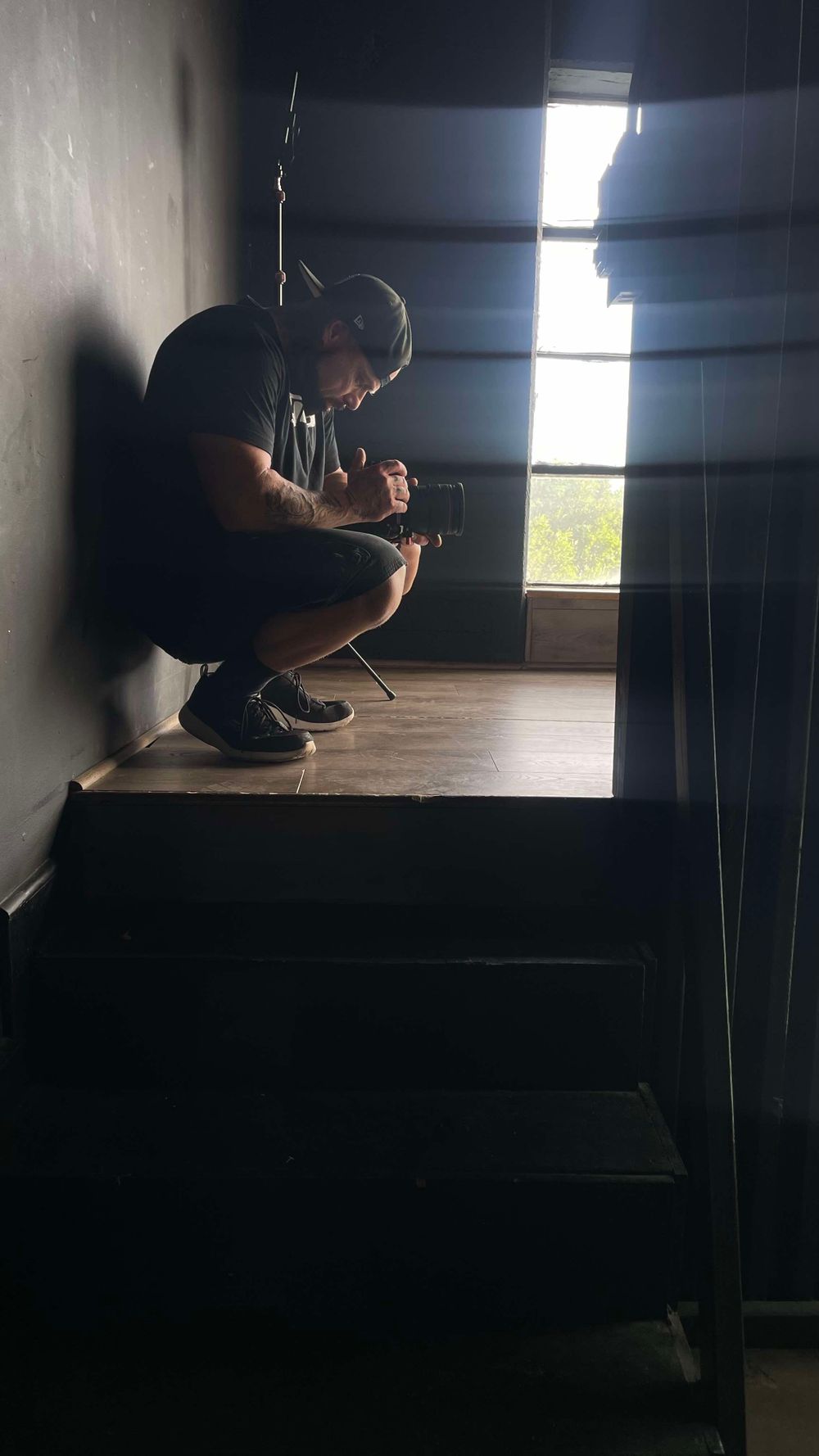 A silhouetted figure crouches by a sunlit window in a dark room while looking at something in their hands.