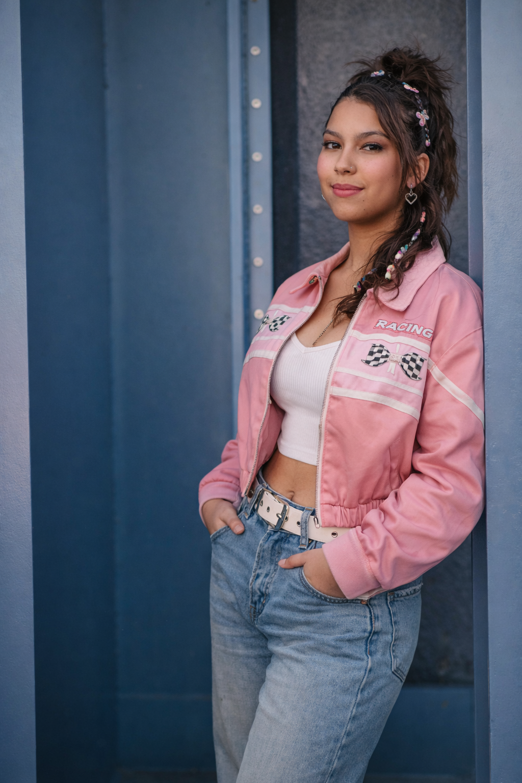 A person poses in a pink cropped jacket and light wash jeans against a blue industrial background.