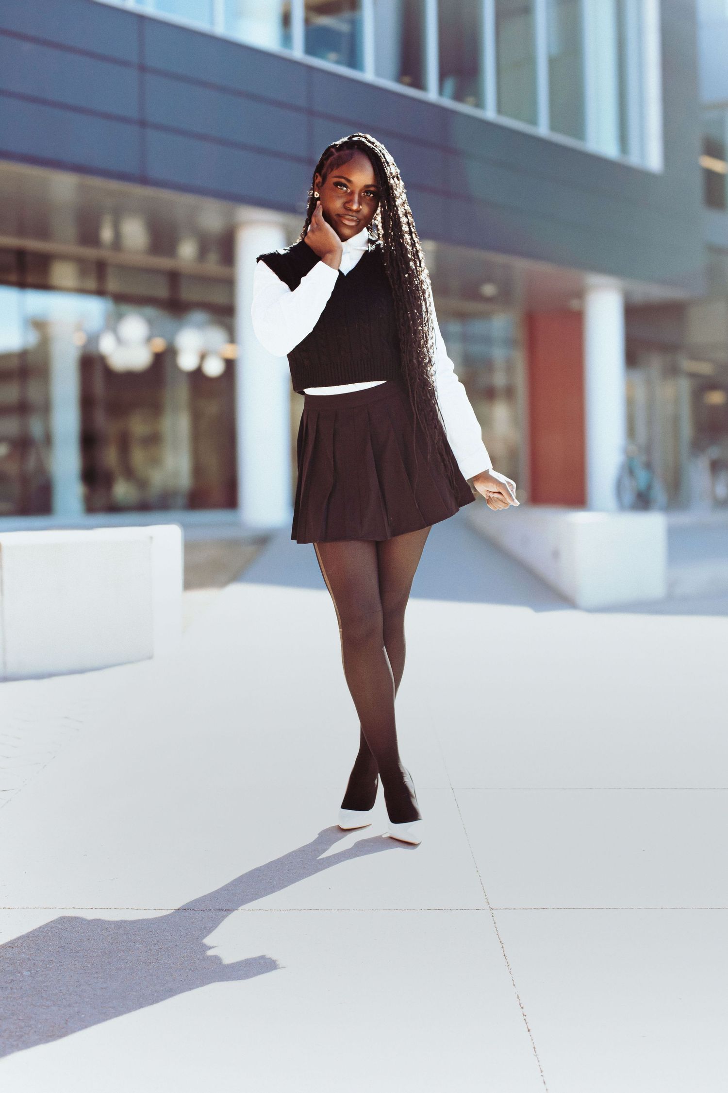 A student in a black pleated skirt and white collared top poses gracefully on a bright urban sidewalk.