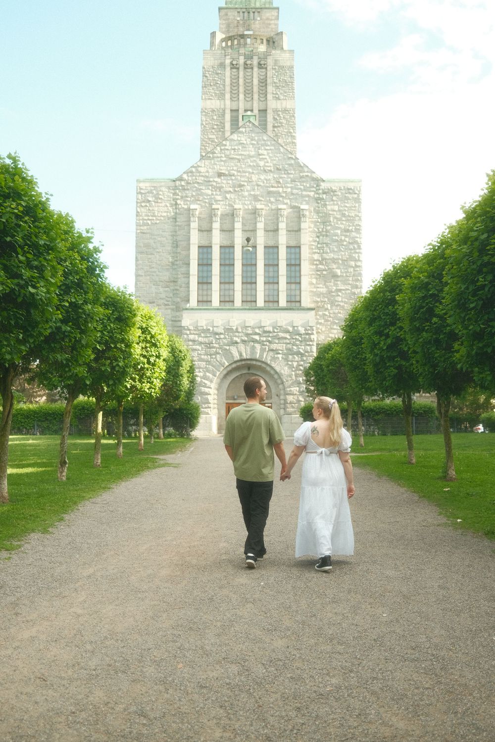 A couple walks along a tree-lined path leading to a grand stone church with a tall tower.