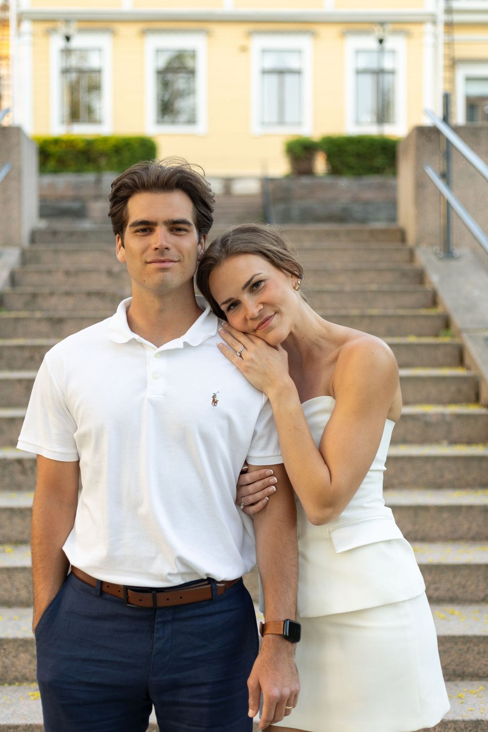 Elegant couple posing on stone steps with yellow building in background.