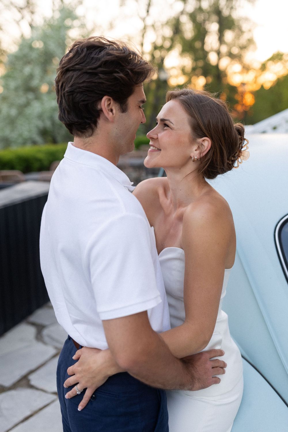 A couple in white attire embraces by a vintage car during sunset.