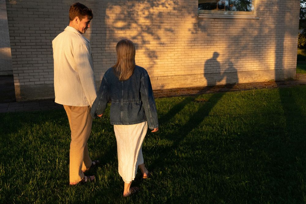 Two people stand together casting long shadows on grass at sunset against a brick wall.