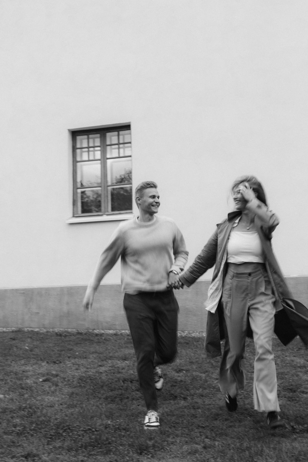 A couple runs and laughs together on a grassy lawn beside a white building with a window.
