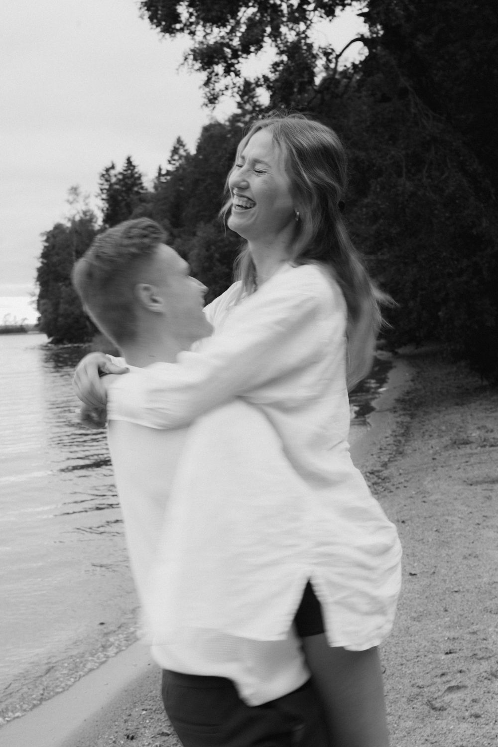 A joyful embracing couple shares a lighthearted moment on a beach with trees in the background.