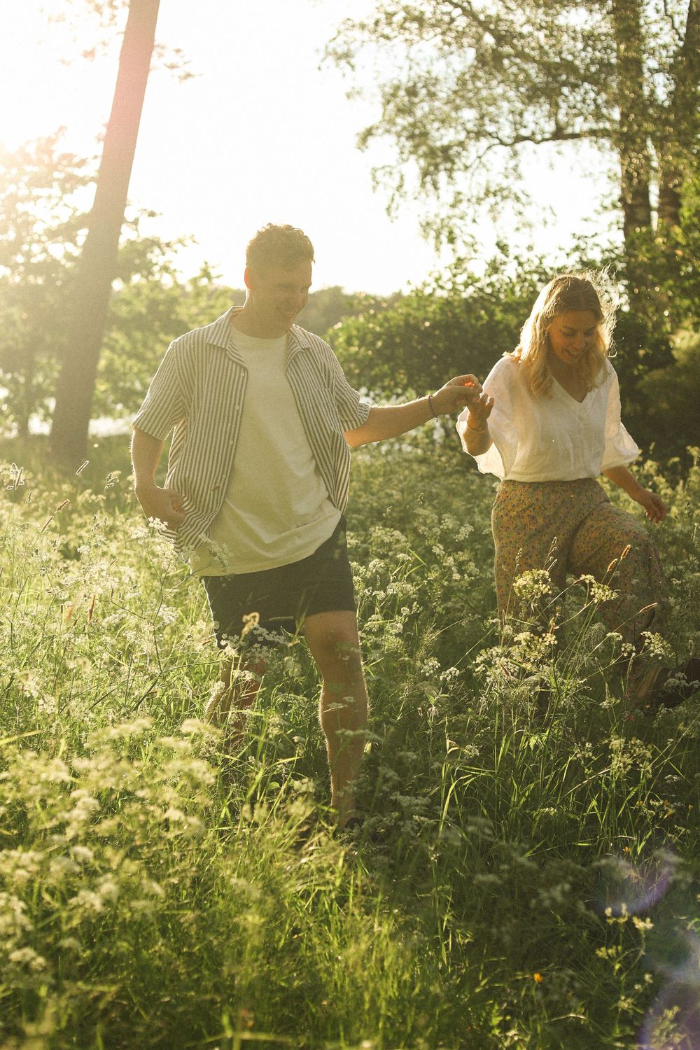 A couple walks together through a sunlit meadow of tall grass during golden hour.